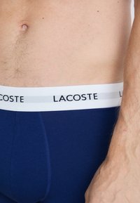 Boxers bleu marine avec une ceinture blanche portant "LACOSTE" imprimé en noir. Texture lisse, coupe ajustée et détails de couture contrastants.
