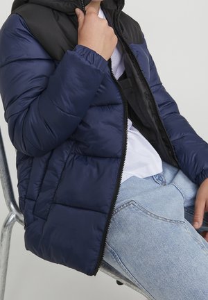 Navy blue puffer jacket with a black hood, featuring a front zipper, textured quilted design, and a relaxed fit. Worn over a white shirt.
