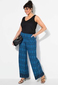 Černý bezramínkový top spojený s modrými kalhotami s širokými nohavicemi a geometrickým vzorem. Modelka drží černou kabelku s hladkým povrchem.