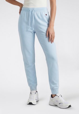 Persona con pantalones joggers azul claro con cordón y zapatillas blancas con suelas negras, de pie con una mano apoyada en la cadera.