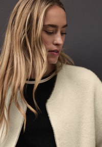 Jasno-kremowy kardigan o miękkiej, fakturowanej powierzchni, z okrągłym kołnierzem i otwartym przodem, noszony na czarnej bluzce z paskowanym dekoltem.