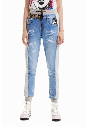 Schmal geschnittene Jogginghose mit Denim-Frontpartien, die weißen Textdruck zeigen, grauen Stoffseitenpartien, einem schwarzen gemusterten Gürtel und weißen Plateausneakern.