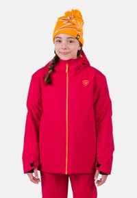 Helle pinke wasserdichte Jacke mit orangefarbendem Reißverschluss, Kapuze und Logo. Weiche Textur mit verstärkten Bündchen. Getragen mit einer bunten orangefarbenen Strickmütze.