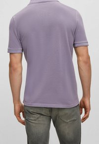 Polo di colore viola chiaro con una finitura testurizzata e un colletto a maglia piatta, caratterizzata da maniche corte e orlo dritto. Inquadrata da dietro.