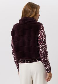 Korte paarse nepbontvest met een gestructureerd golvend patroon, gedragen over een blouse met lange mouwen en patroon. Onderaan zichtbaar crèmekleurige broek.