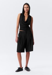 Black sleeveless top with a cowl neck and hood, paired with high-waisted black pleated shorts. Worn with black lace-up shoes.