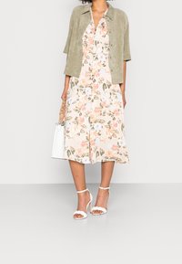 Robe midi à fleurs de couleur claire avec manches courtes, associée à une veste en daim beige. Talons blancs à brides et un sac à main blanc complètent le look.