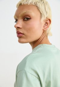 T-shirt in cotone verde menta con collo a giro, texture morbida e maniche corte, indossato da una persona con capelli biondo platino e piercings visibili.