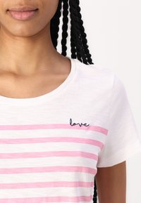 Hvit t-skjorte med rosa horisontale striper og et marineblått brodert "love" på venstre bryst. Mykt stoff med en avslappet passform og rund hals.