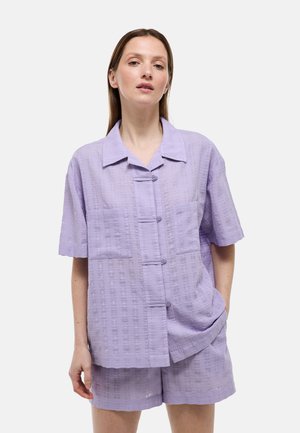 Femme portant une chemise lavande ample à manches courtes avec deux poches avant et un short assorti, debout avec une main dans la poche.