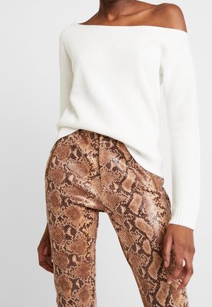 Vrouw draagt een witte ribgebreide trui met blote schouders en een hooggesneden bruine broek met slangenprint, staand met handen ontspannen langs het lichaam.