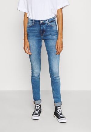 Personne portant un jean skinny bleu retroussé, des baskets montantes noires et blanches, et une chemise blanche à manches courtes, debout devant un fond uni.