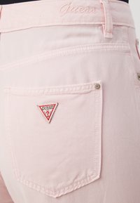 Ροζ τζιν παντελόνι ελαφρού χρώματος, που διαθέτει ένα τριγωνικό λογότυπο Guess σε κόκκινο και λευκό χρώμα, ραμμένο στην πίσω τσέπη, με διακριτές ραφές και μεταλλικά σκουλαρίκια.
