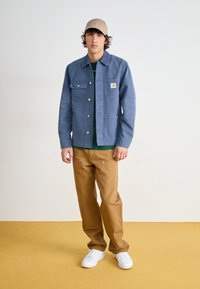 Lyseblå denimjakke med to brystlommer og gullknapper, lagd over en grønn skjorte, kombinert med brune bukser og hvite joggesko.