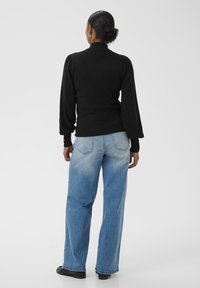 Femeie cu părul închis la culoare, coafat în coc, îmbrăcată cu o bluză neagră cu mâneci lungi și blugi largi de culoare bleu deschis, stând cu spatele pe un fundal simplu.