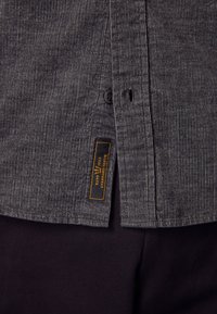 Tessuto della camicia a righe grigie con chiusura a bottoni e etichetta laterale che mostra "Standard Issue" in arancione. Tessuto scuro sotto, a enfatizzare il contrasto.