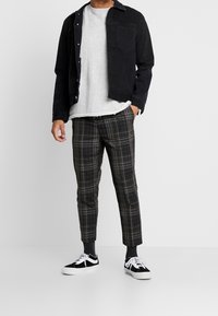 Uomo che indossa una giacca di jeans nera sopra una camicia bianca, pantaloni a quadri grigi e neri, calze nere e sneaker bianche e nere, in piedi contro uno sfondo bianco.