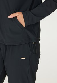 Top sportivo nero con tasca frontale e pantaloni abbinati. Presenta un tessuto texturizzato, un zip decorativo e un accento dorato con logo.