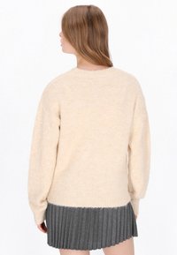 Cremfarbener Strickpullover mit weicher Textur, Ballonärmeln und einem grauen, plissierten Rock am unteren Saum. Keine sichtbaren Muster oder Hardware.