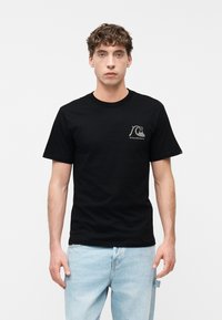 Νεαρός άνδρας που φοράει μαύρο t-shirt με λευκό λογότυπο κύματος στο στήθος και ανοιχτό μπλε τζιν, στέκεται μπροστά σε ένα απλό λευκό φόντο.
