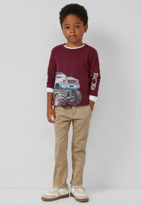 Rotweinfarbenes Langarmshirt mit blau-weißer Monster-Truck-Grafik, kombiniert mit beigen Hosen und weißen Sneakers mit braunen Sohlen.