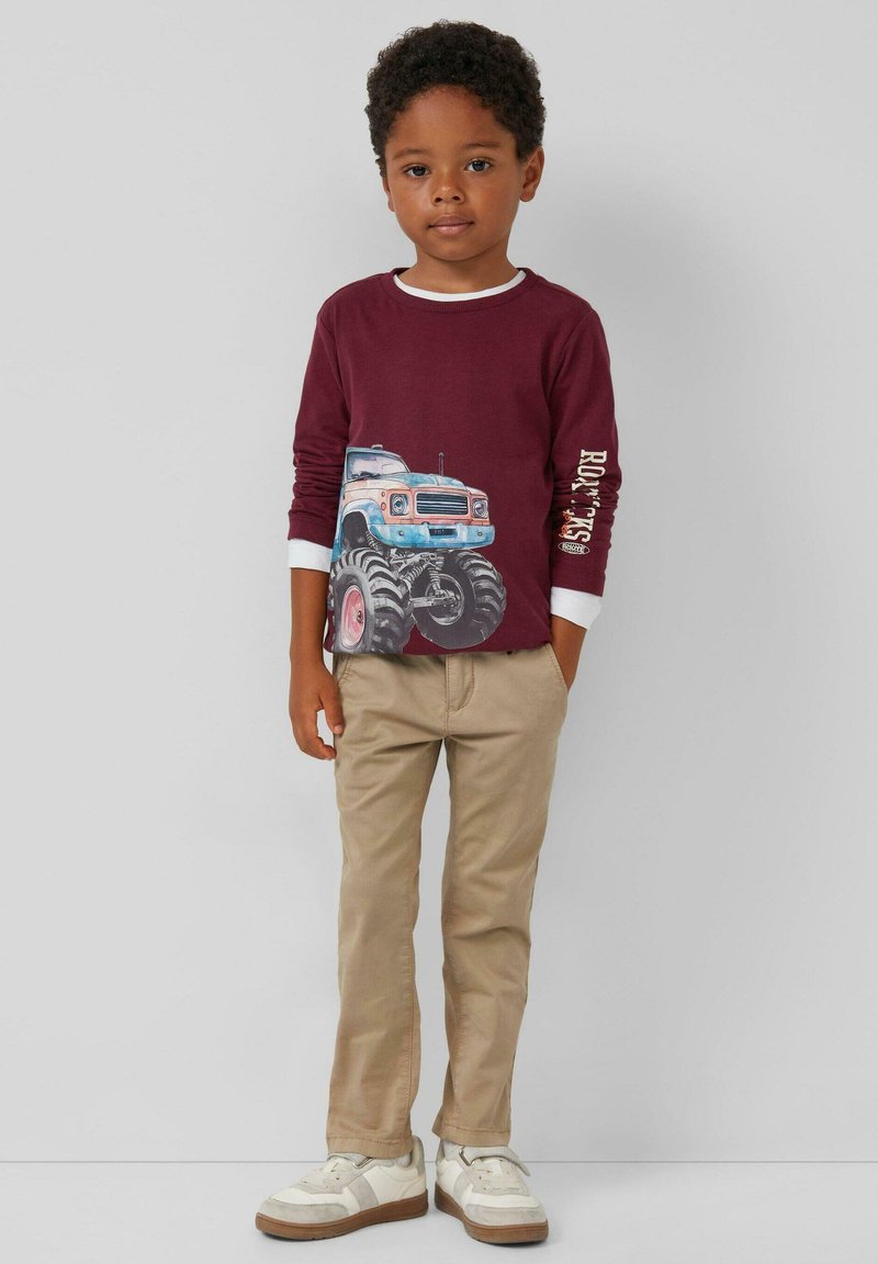 Rotweinfarbenes Langarmshirt mit blau-weißer Monster-Truck-Grafik, kombiniert mit beigen Hosen und weißen Sneakers mit braunen Sohlen.