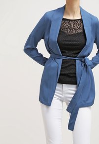 Blauwe satijnen wikkeljack met lange mouwen en een gestrikte taille, gedragen over een zwart met patroon top, gecombineerd met witte broeken.
