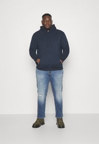 Mørkeblå hettegenser med frontlomme, kombinert med lyseblå ripped jeans og mørkegrønne snørestøvler. Ensfarget hvit bakgrunn.
