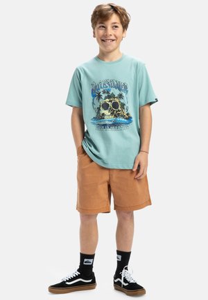 Turkis bomulds t-shirt med et grafisk print af et kranium, palmetræer og bølger; parret med brune shorts og sorte sneakers.