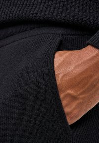 Czarny dzianinowy sweter zbliżenie ukazujące fakturowaną tkaninę oraz boczną kieszeń. Ręka swobodnie opiera się o tkaninę, podkreślając splot.