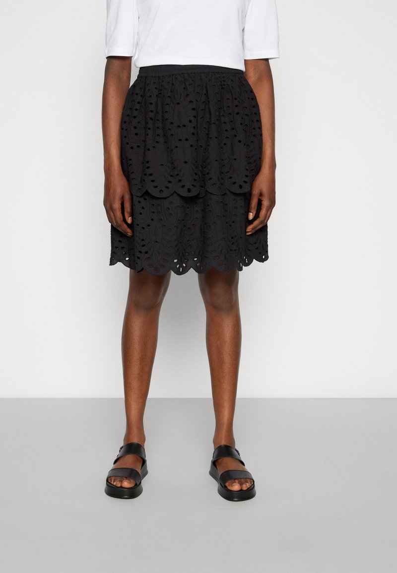 Jupe en dentelle noire à œillets avec un design superposé, dotée d'un ourlet festonné et d'une taille élastique, associée à des sandales plates noires.
