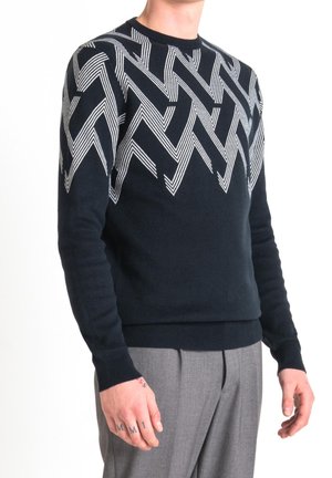 Homme portant un pull noir avec un motif de lignes en zigzag blanches sur les épaules et le haut de la poitrine, associé à un pantalon gris.