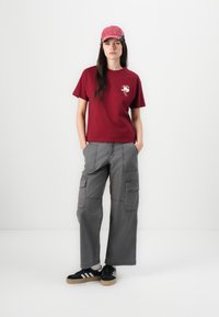 Bordeaux bomulls t-skjorte med grafisk design, grå cargo-bukser med lommer, og svarte sneakers med hvite striper og gummisåler. Rosa caps.
