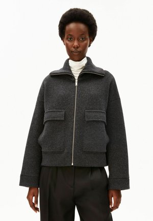 Frau mit kurzem, natürlichem Haar, die eine dunkelgraue Jacke mit Reißverschluss und Klappentaschen sowie eine schwarze Hose trägt, vor weißem Hintergrund.