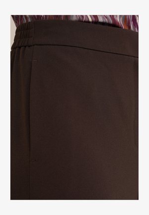 Brązowe, gładkie spodnie z plisowanym pasem i bocznymi szwami. Zbliżenie podkreślające układ materiału i szczegóły wzornictwa.