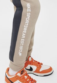 Beige sweatpants met een donkerblauwe zijstreep, met witte branding die "NIKE SPORTSWEAR EST. 1972." zegt. Gecombineerd met oranje en witte sneakers.