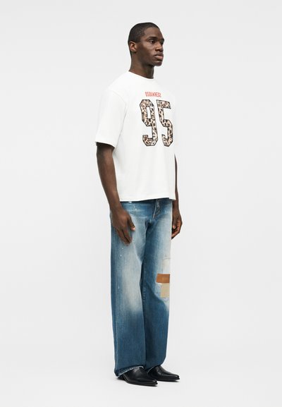 Άνδρας που στέκεται φορώντας λευκό μπλουζάκι με το "95" σε λεοπάρ τύπωμα και μπλε τζιν με λεπτομέρειες σε μπαλώματα και μαύρα μυτερά παπούτσια.