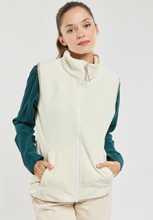 Femme aux cheveux bruns attachés porte une veste crème zippée sur un haut vert à manches longues, les mains dans les poches de la veste, debout devant un fond blanc.