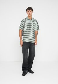 Striped polo shirt in light green with black lines, paired with dark baggy jeans and black shoes. The model carries a gray shoulder bag.