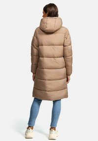 Long beige padded coat with a hood, featuring horizontal quilting. Paired with blue jeans and light-colored sneakers. Smooth texture.