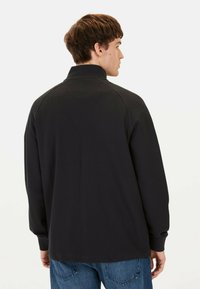 Zwarte poloshirt met lange mouwen en hoge kraag, van een gestructureerde stof met ribbelmanchetten. De achterkant heeft een gladde afwerking en een ontspannen pasvorm.