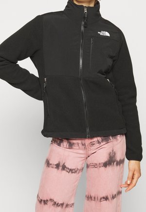 Person iført en sort fleecejakke med lynlås og The North Face-logo samt lyserøde bukser med batikmønster, stående med den ene hånd på hoften.