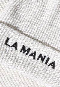 Bílá pletená čepice s ribovým vzorem a ohrnutým lemem, s výraznou černou výšivkou "LA MANIA" na přední části. Měkká textura s těsným střihem.
