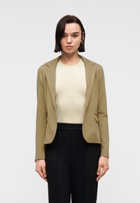 Jeune personne aux cheveux courts et fonc és, portant un blazer beige, un haut crème et un pantalon noir, debout devant un fond clair uni.