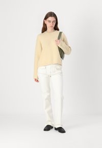 Beige textuurtrui met lange mouwen, gecombineerd met witte rechte spijkerbroek en zwarte schoenen. Model houdt een olijfgroene tas vast.