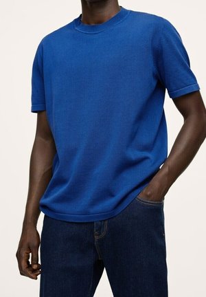 Hombre que lleva una camiseta de punto azul lisa de manga corta y vaqueros azul oscuro, con una mano en el bolsillo y la otra relajada a su lado.