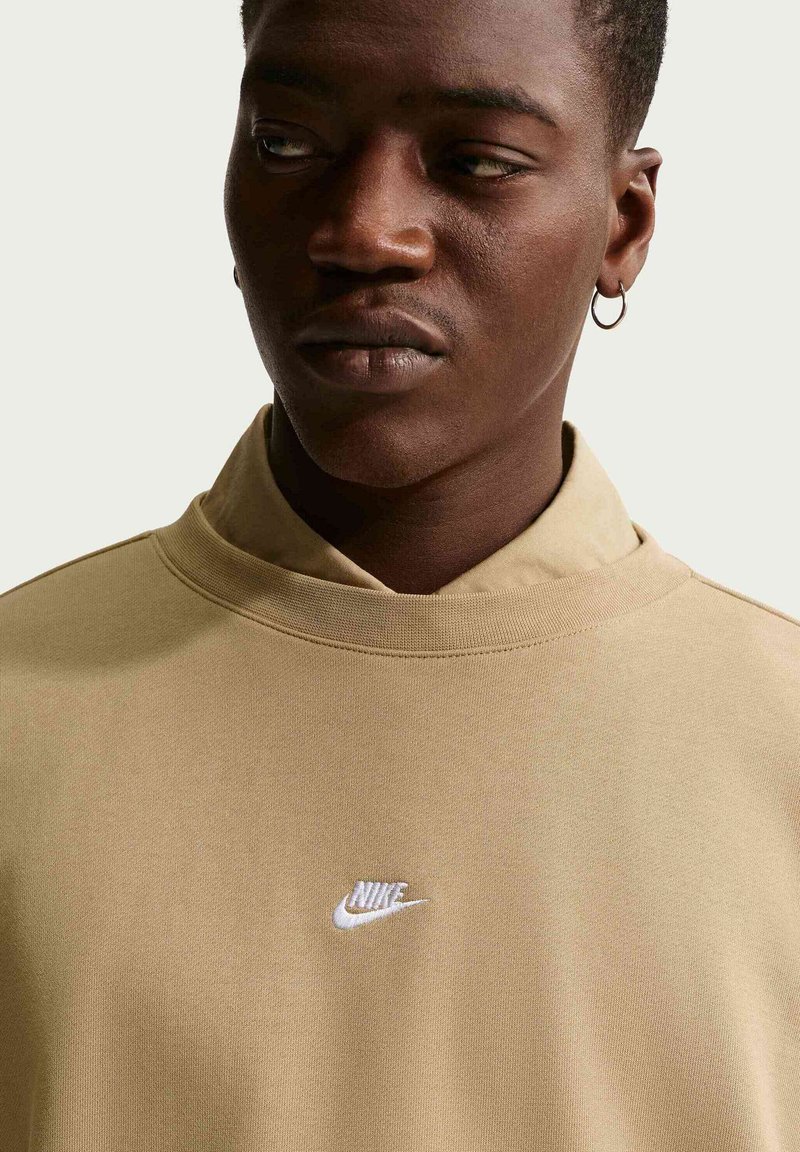 Giovane uomo che indossa una felpa Nike beige con un piccolo logo bianco, orecchino ad anello d’argento, che guarda di lato su uno sfondo chiaro e uniforme.
