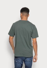 T-shirt in cotone verde scuro con collo a giro, maniche corte e vestibilità rilassata; la vista posteriore mostra una superficie liscia e un orlo cucito.