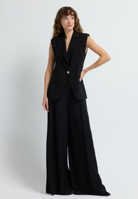 Blazer senza maniche nero con scollatura a V, bottone a forma di cuore dorato e tasche a pattina, abbinato a pantaloni neri a gamba larga. Tessuto dalla superficie liscia.