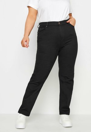 Persoon die zwarte jeans met rechte pijpen draagt, witte plateau sneakers en een wit T-shirt met korte mouwen, staand met één hand in de zak.